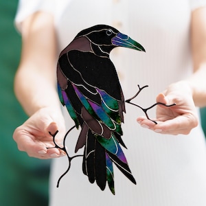 Peut inclure: Une sculpture de corbeau en vitrail avec un corps noir, des ailes irisées violettes et sarcelle, et un bec bleu-vert. Le corbeau est perché sur une branche en métal foncé. L'œuvre est présentée sur fond blanc.