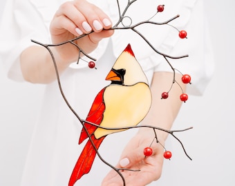 May include: A stained glass cardinal bird with red, yellow, and black details perched on a metal branch with red berries. The bird is a decorative piece, likely for a window or wall.