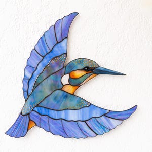 K&ouml;nnte beinhalten: Ein Buntglasbild eines fliegenden Eisvogels. Der Vogel hat Blaut&ouml;ne, Lila und Orange, mit einem langen, spitzen Schnabel. Die Fl&uuml;gel sind weit ausgebreitet und zeigen die komplizierten Details der Glasarbeiten.