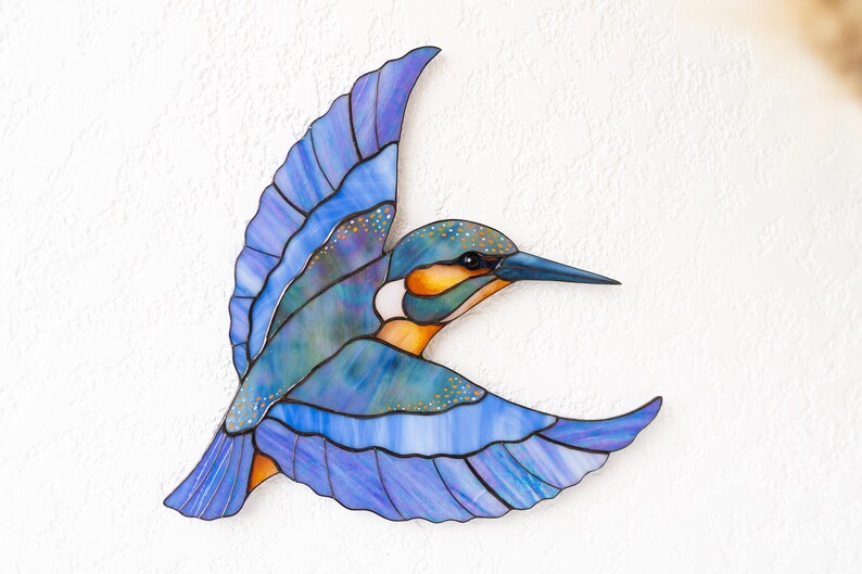 K&ouml;nnte beinhalten: Ein Buntglasbild eines fliegenden Eisvogels. Der Vogel hat Blaut&ouml;ne, Lila und Orange, mit einem langen, spitzen Schnabel. Die Fl&uuml;gel sind weit ausgebreitet und zeigen die komplizierten Details der Glasarbeiten.