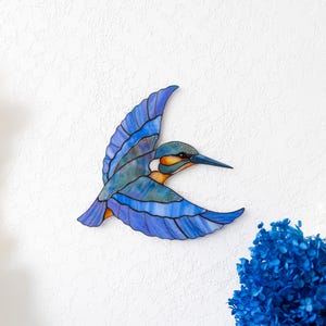K&ouml;nnte beinhalten: Ein fliegender Eisvogel aus Buntglas, mit Blau-, T&uuml;rkis- und Oranget&ouml;nen. Der Vogel ist an einer wei&szlig;en Wand befestigt, mit einem Strau&szlig; blauer Blumen in der unteren rechten Ecke.