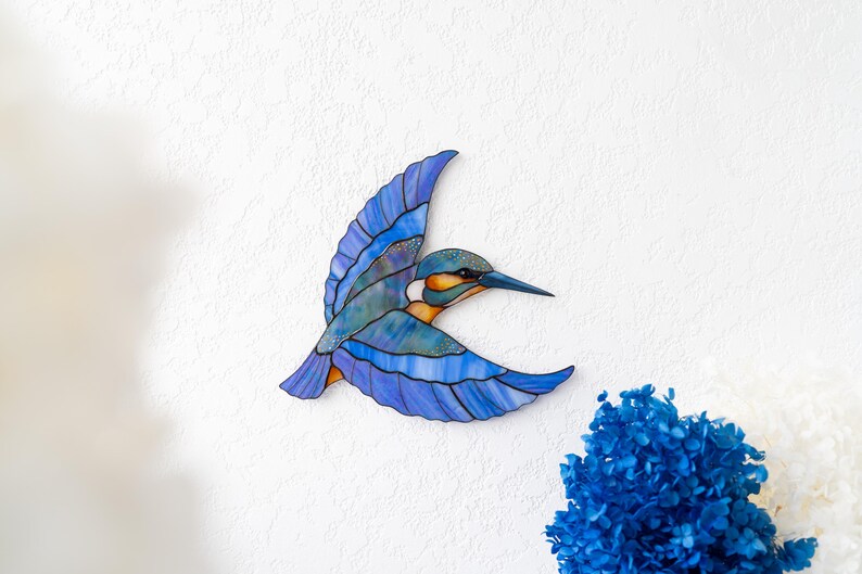K&ouml;nnte beinhalten: Ein fliegender Eisvogel aus Buntglas, mit Blau-, T&uuml;rkis- und Oranget&ouml;nen. Der Vogel ist an einer wei&szlig;en Wand befestigt, mit einem Strau&szlig; blauer Blumen in der unteren rechten Ecke.