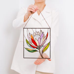 Puede incluir: Panel de vidriera con un dise&ntilde;o de flor de protea colorida. La flor tiene p&eacute;talos en tonos de rosa, rojo, naranja y blanco, con hojas verdes y un tallo marr&oacute;n. El panel est&aacute; enmarcado en metal oscuro y cuelga de una cadena.