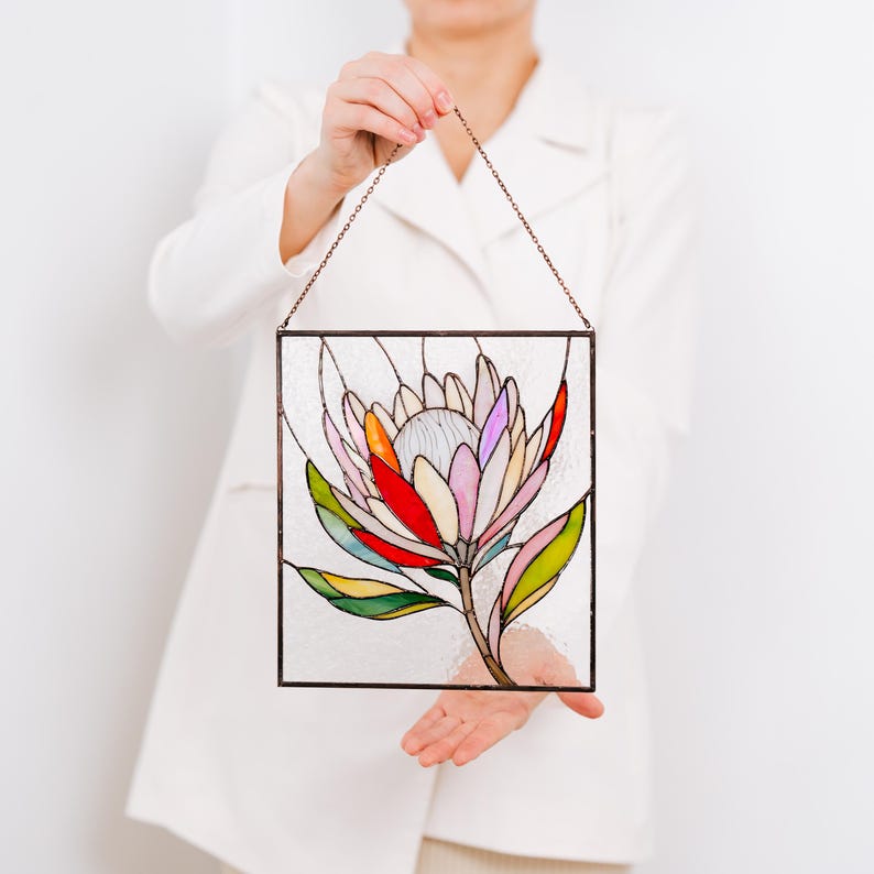 Puede incluir: Panel de vidriera con un dise&ntilde;o de flor de protea colorida. La flor tiene p&eacute;talos en tonos de rosa, rojo, naranja y blanco, con hojas verdes y un tallo marr&oacute;n. El panel est&aacute; enmarcado en metal oscuro y cuelga de una cadena.