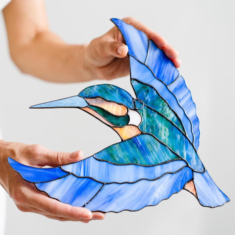 K&ouml;nnte beinhalten: Eine Glasmalerei-Vogelskulptur im Flug, mit Blaut&ouml;nen, T&uuml;rkis und Wei&szlig;. Der Vogel hat einen langen Schnabel und ausgebreitete Fl&uuml;gel, mit einem kleinen Metallring zum Aufh&auml;ngen. Das Kunstwerk wird von H&auml;nden vor wei&szlig;em Hintergrund gehalten.