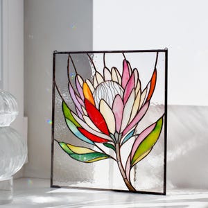 Puede incluir: Panel de vidriera con un dise&ntilde;o de flor de protea colorida. La flor tiene p&eacute;talos en tonos de rosa, rojo, naranja y amarillo, con hojas verdes y un centro blanco. El panel est&aacute; enmarcado en negro y est&aacute; dise&ntilde;ado para colgar.