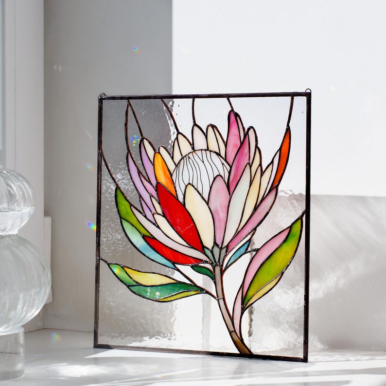 Puede incluir: Panel de vidriera con un dise&ntilde;o de flor de protea colorida. La flor tiene p&eacute;talos en tonos de rosa, rojo, naranja y amarillo, con hojas verdes y un centro blanco. El panel est&aacute; enmarcado en negro y est&aacute; dise&ntilde;ado para colgar.