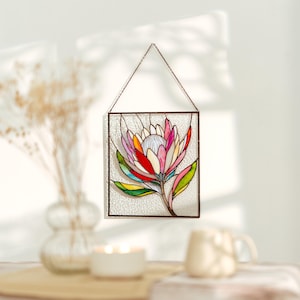 Puede incluir: Un panel de vidrieras con un dise&ntilde;o de flor de protea colorida. La flor tiene p&eacute;talos rosas, rojos, amarillos y verdes, con un centro blanco. El panel est&aacute; enmarcado en metal oscuro y cuelga de una cadena.