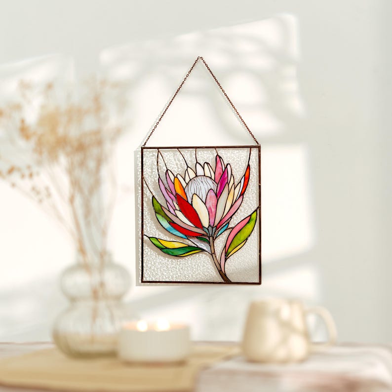 Puede incluir: Un panel de vidrieras con un dise&ntilde;o de flor de protea colorida. La flor tiene p&eacute;talos rosas, rojos, amarillos y verdes, con un centro blanco. El panel est&aacute; enmarcado en metal oscuro y cuelga de una cadena.