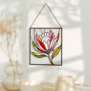 Puede incluir: Panel de vidrieras con un dise&ntilde;o de flor de protea colorida. La flor tiene p&eacute;talos en tonos rosa, rojo, amarillo y naranja, con hojas verdes. El panel est&aacute; enmarcado en metal oscuro y cuelga de una cadena.