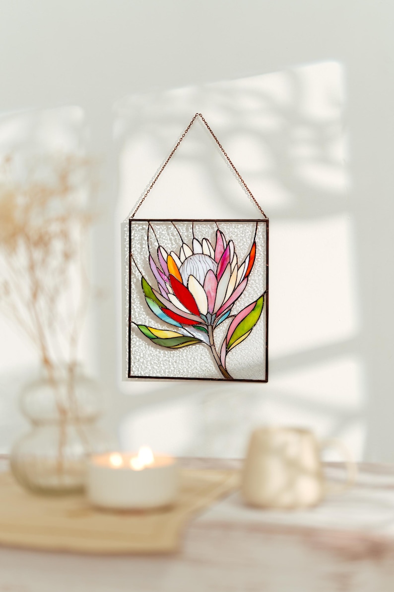 Puede incluir: Panel de vidrieras con un dise&ntilde;o de flor de protea colorida. La flor tiene p&eacute;talos en tonos rosa, rojo, amarillo y naranja, con hojas verdes. El panel est&aacute; enmarcado en metal oscuro y cuelga de una cadena.