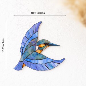 K&ouml;nnte beinhalten: Ein Buntglas-Eisvogel im Flug, mit blauen, orangefarbenen und wei&szlig;en Farbt&ouml;nen. Das Kunstwerk ist 26 cm hoch und 26 cm breit. Die Fl&uuml;gel des Vogels sind ausgebreitet und sein Schnabel ist nach vorne gerichtet.