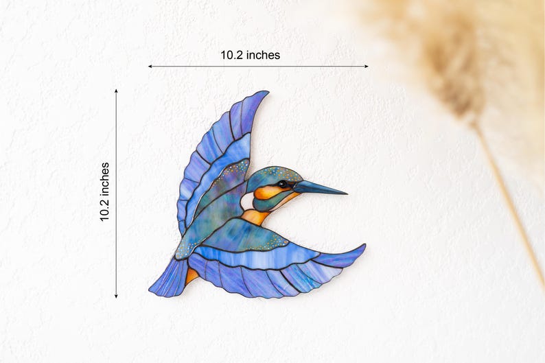 K&ouml;nnte beinhalten: Ein Buntglas-Eisvogel im Flug, mit blauen, orangefarbenen und wei&szlig;en Farbt&ouml;nen. Das Kunstwerk ist 26 cm hoch und 26 cm breit. Die Fl&uuml;gel des Vogels sind ausgebreitet und sein Schnabel ist nach vorne gerichtet.