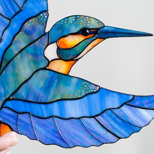 K&ouml;nnte beinhalten: Glasmalerei eines Eisvogels in Blau-, Orange- und Wei&szlig;t&ouml;nen. Der Vogel ist im Flug dargestellt, mit detaillierten Fl&uuml;geln und einer strukturierten Oberfl&auml;che. Die Konturen sind schwarz.