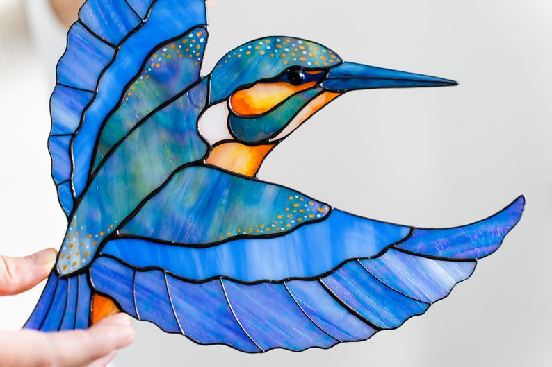 K&ouml;nnte beinhalten: Glasmalerei eines Eisvogels in Blau-, Orange- und Wei&szlig;t&ouml;nen. Der Vogel ist im Flug dargestellt, mit detaillierten Fl&uuml;geln und einer strukturierten Oberfl&auml;che. Die Konturen sind schwarz.