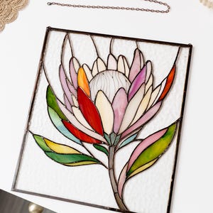 Puede incluir: Vidriera con un dise&ntilde;o de flor de protea colorida. La flor tiene p&eacute;talos en tonos rosa, rojo, naranja y blanco, con hojas verdes. La obra de arte est&aacute; enmarcada en metal oscuro e incluye una cadena para colgar.