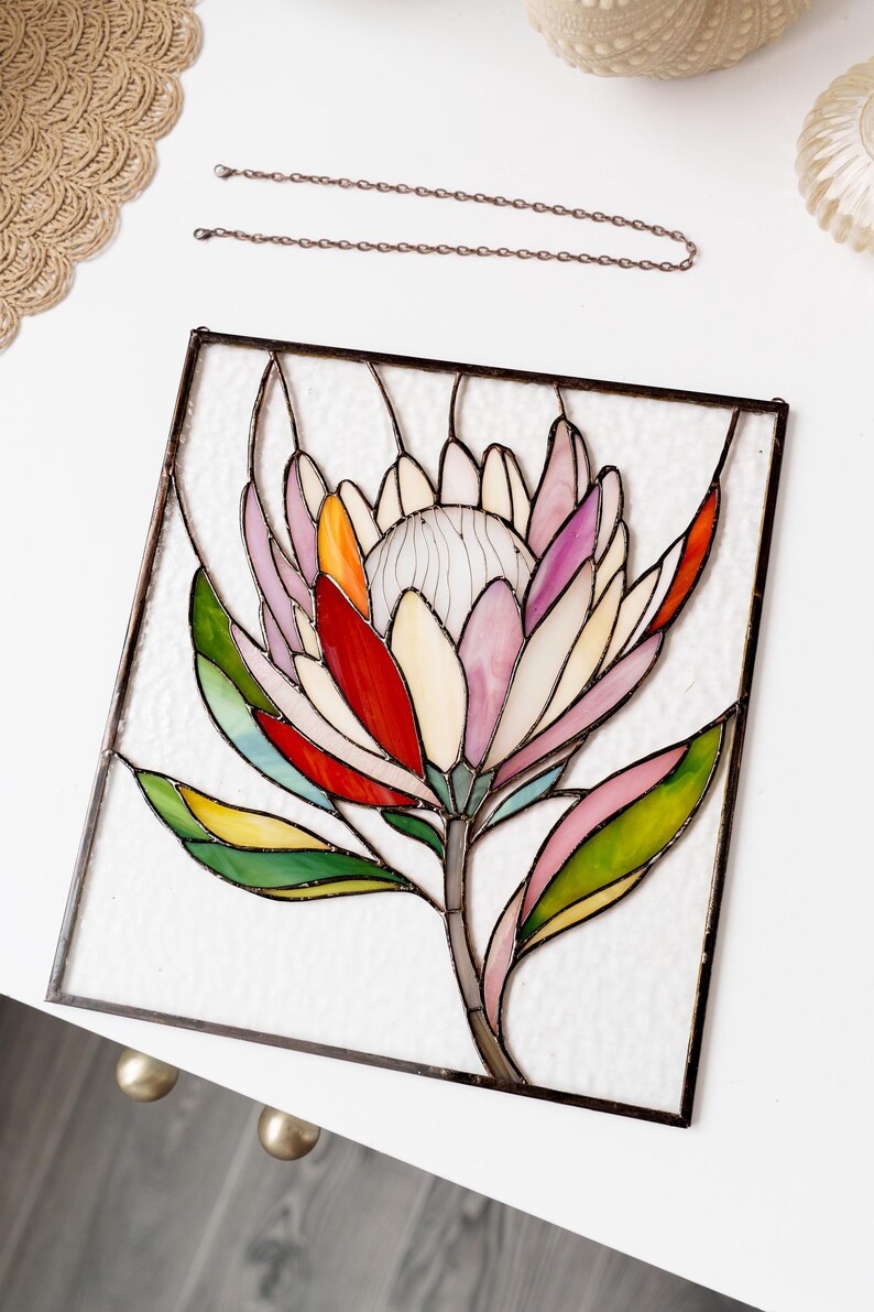 Puede incluir: Vidriera con un dise&ntilde;o de flor de protea colorida. La flor tiene p&eacute;talos en tonos rosa, rojo, naranja y blanco, con hojas verdes. La obra de arte est&aacute; enmarcada en metal oscuro e incluye una cadena para colgar.