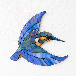 K&ouml;nnte beinhalten: Ein Buntglas-Eisvogel im Flug, mit Blaut&ouml;nen, T&uuml;rkis, Orange und Gelb. Der Vogel hat einen langen, spitzen Schnabel und detaillierte Fl&uuml;gelmuster. Das Kunstwerk ist an einer wei&szlig;en, strukturierten Wand befestigt.