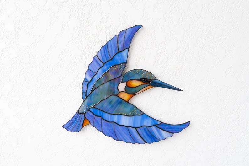 K&ouml;nnte beinhalten: Ein Buntglas-Eisvogel im Flug, mit Blaut&ouml;nen, T&uuml;rkis, Orange und Gelb. Der Vogel hat einen langen, spitzen Schnabel und detaillierte Fl&uuml;gelmuster. Das Kunstwerk ist an einer wei&szlig;en, strukturierten Wand befestigt.