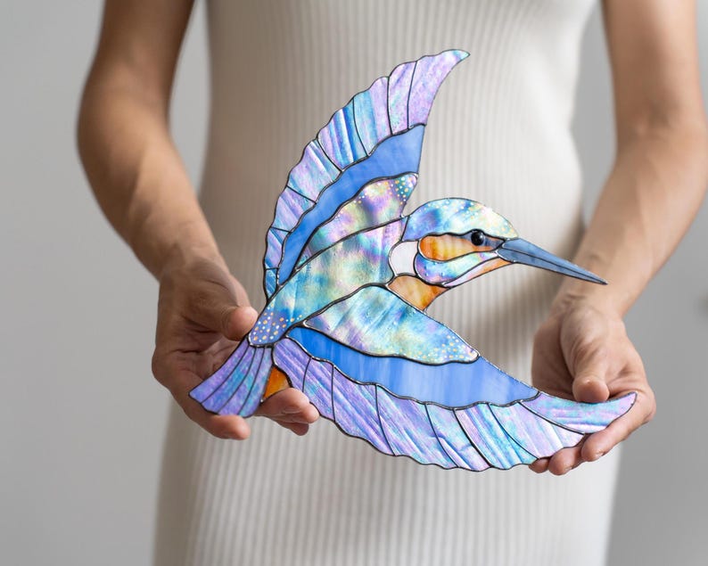 K&ouml;nnte beinhalten: Glasmalerei-Skulptur eines Eisvogels im Flug. Das Kunstwerk hat blaue, violette und schillernde Fl&uuml;gel mit einer orangefarbenen und wei&szlig;en Brust. Der Schnabel ist lang und spitz. Die Skulptur wird vor einem wei&szlig;en Hintergrund gehalten. Die Fl&uuml;gelspannweite betr&auml;gt ca. 25 cm.