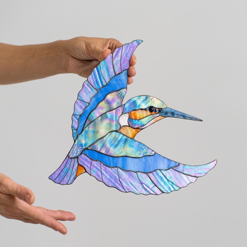 K&ouml;nnte beinhalten: Ein Buntglasvogel im Flug, in Blau-, Lila- und Oranget&ouml;nen. Die Fl&uuml;gel des Vogels sind ausgebreitet und zeigen detaillierte Muster und schillernde Effekte. Das Kunstwerk wird von einer Hand gehalten, was die Handwerkskunst hervorhebt.