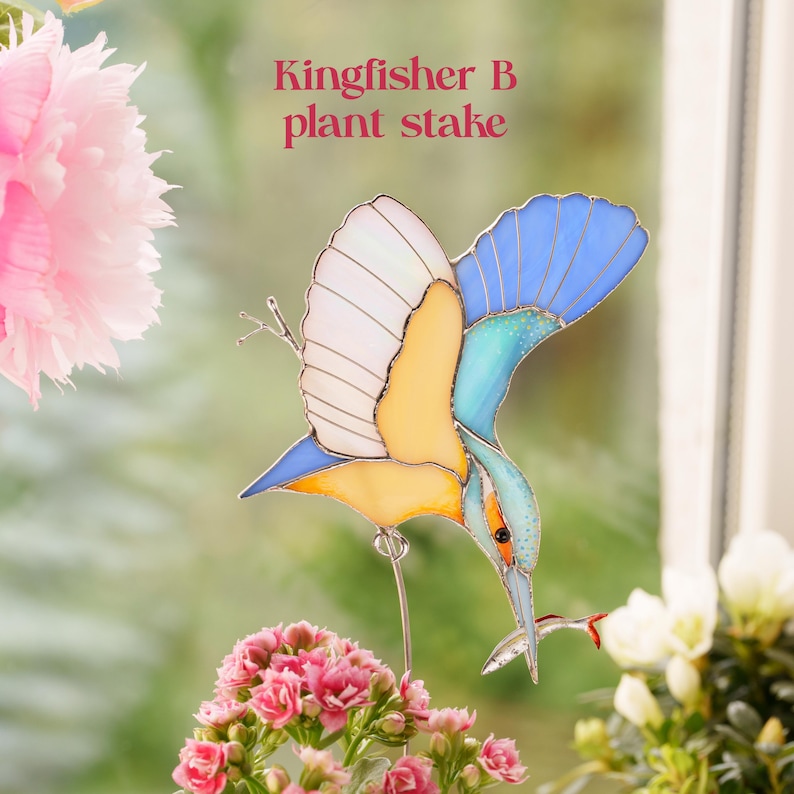 Pu&ograve; includere: Un tutore per piante in vetro colorato a forma di martin pescatore in volo, con pannelli di vetro blu, giallo e bianco e saldature in metallo scuro. L'uccello tiene un piccolo pesce nel becco. Il testo Kingfisher B plant stake &egrave; visibile.