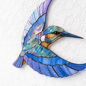 K&ouml;nnte beinhalten: Glasmalerei eines Eisvogels im Flug. Das Kunstwerk zeigt Blaut&ouml;ne, Lila und Orange mit schillernden Akzenten. Das Kunstwerk ist an einer strukturierten wei&szlig;en Wand befestigt.