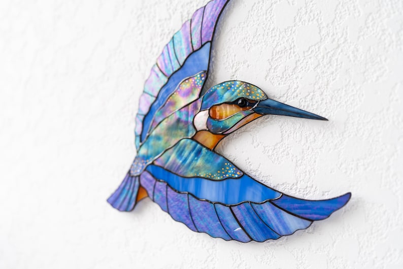 K&ouml;nnte beinhalten: Glasmalerei eines Eisvogels im Flug. Das Kunstwerk zeigt Blaut&ouml;ne, Lila und Orange mit schillernden Akzenten. Das Kunstwerk ist an einer strukturierten wei&szlig;en Wand befestigt.