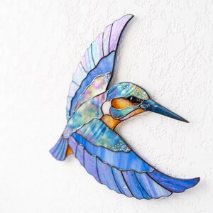 K&ouml;nnte beinhalten: Eine farbenfrohe Vogelskulptur aus Glas in Flugpose. Die Fl&uuml;gel zeigen Blaut&ouml;ne, Lila und schillernde Akzente. Der Schnabel ist dunkelblau, der K&ouml;rper hat orange und gr&uuml;ne Details. Eine dekorative Wanddekoration.