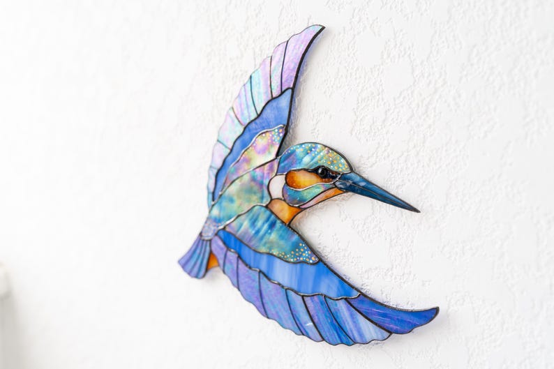 K&ouml;nnte beinhalten: Eine farbenfrohe Vogelskulptur aus Glas in Flugpose. Die Fl&uuml;gel zeigen Blaut&ouml;ne, Lila und schillernde Akzente. Der Schnabel ist dunkelblau, der K&ouml;rper hat orange und gr&uuml;ne Details. Eine dekorative Wanddekoration.