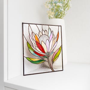 Puede incluir: Panel de vidriera con una flor de protea colorida. La flor tiene p&eacute;talos en tonos rosa, naranja, rojo y amarillo, con hojas verdes y un tallo marr&oacute;n. La obra de arte est&aacute; enmarcada en metal oscuro y se encuentra sobre una superficie blanca.