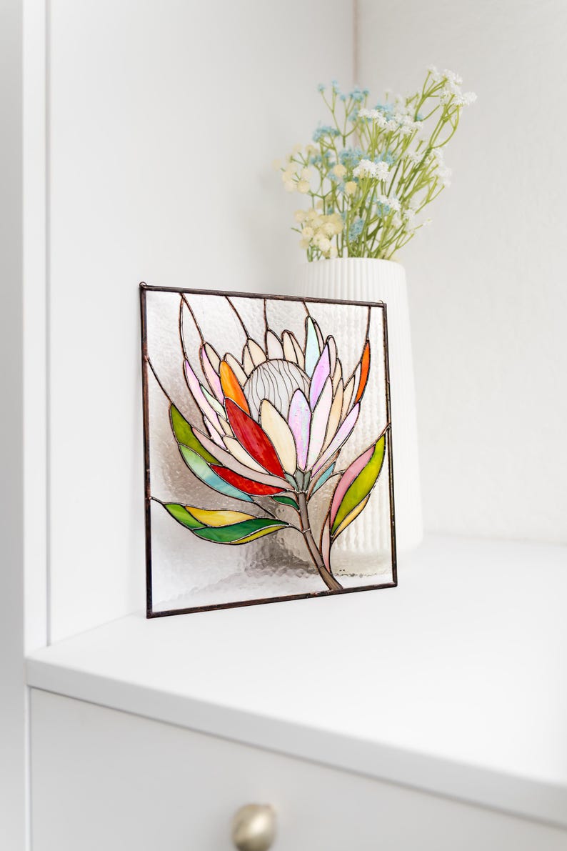 Puede incluir: Panel de vidriera con una flor de protea colorida. La flor tiene p&eacute;talos en tonos rosa, naranja, rojo y amarillo, con hojas verdes y un tallo marr&oacute;n. La obra de arte est&aacute; enmarcada en metal oscuro y se encuentra sobre una superficie blanca.