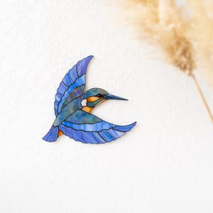 K&ouml;nnte beinhalten: Ein Buntglasbild eines Eisvogels im Flug. Der Vogel hat blaue, gr&uuml;ne und orange Farbt&ouml;ne und ausgebreitete Fl&uuml;gel. Das Kunstwerk ist an einer wei&szlig;en Wand befestigt, mit getrocknetem Ziergras im Hintergrund.