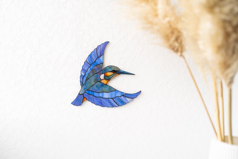 K&ouml;nnte beinhalten: Ein Buntglasbild eines Eisvogels im Flug. Der Vogel hat blaue, gr&uuml;ne und orange Farbt&ouml;ne und ausgebreitete Fl&uuml;gel. Das Kunstwerk ist an einer wei&szlig;en Wand befestigt, mit getrocknetem Ziergras im Hintergrund.