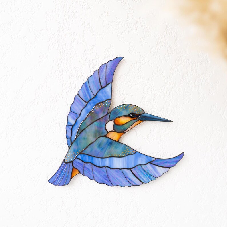 K&ouml;nnte beinhalten: Ein Buntglas-Eisvogel im Flug, mit Blaut&ouml;nen, Lila und Orange. Die Fl&uuml;gel des Vogels sind ausgebreitet und zeigen detaillierte Muster und eine strukturierte Oberfl&auml;che. Das Kunstwerk ist an einer wei&szlig;en Wand befestigt.