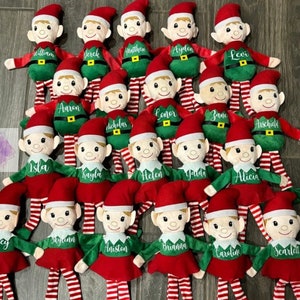 May include: A collection of plush Christmas elves, each with a red hat, green outfit, and white and red striped legs. Each elf has a name embroidered on its outfit. The elves are arranged on a wooden surface.