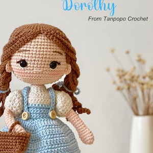 May include: Crocheted doll with brown hair, a blue dress, and red shoes. She is holding a brown basket. The doll is named Dorothy and is from Tanpopo Crochet.
