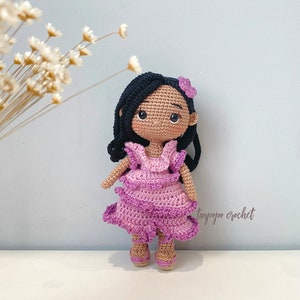 May include: Crocheted doll with long black hair, wearing a pink ruffled dress and brown sandals. The doll has brown eyes and a pink flower in her hair. The doll is standing on a white surface.