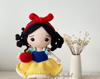 Könnte beinhalten: Eine gehäkelte Schneewittchen-Puppe mit schwarzen Haaren, einer roten Schleife, einem blau-weißen Kleid, einem gelben Rock und einem roten Apfel. Die Puppe steht auf einer weißen Oberfläche.