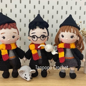 Può includere: Tre personaggi di Harry Potter all'uncinetto, Ron, Harry ed Hermione, che indossano vesti nere, sciarpe gialle e rosse e cappelli neri a punta. I personaggi sono in piedi su una superficie di legno con un topo bianco di fronte a loro. Il testo "Tanpopo Crochet" è visibile nell'immagine.