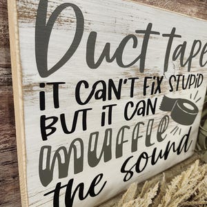 May include: A rectangular wooden sign with a distressed white background and black lettering. The text reads "Duct tape, it can't fix stupid, but it can muffle the sound." A graphic of duct tape is also included.
