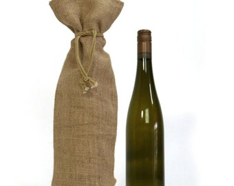 May include: A green wine bottle with a brown cap stands next to a burlap wine bottle bag with a drawstring closure. The bag is made of a rough, textured fabric. The background is white.
