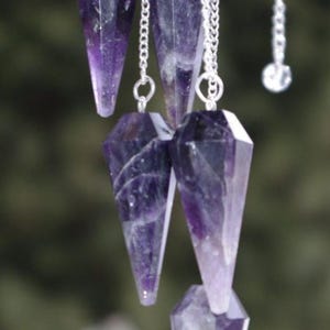 May include: Several amethyst crystal pendulums hang from silver chains. The crystals are faceted, with a pointed bottom and a geometric top. The purple crystals vary in shade, with some having white inclusions.