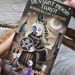 May include: A tarot card deck box with the title "Deviant Moon Tarot Borderless Edition" in gold lettering. The box features a gothic illustration of a figure with a moon face, holding a pentagram, and surrounded by other figures.