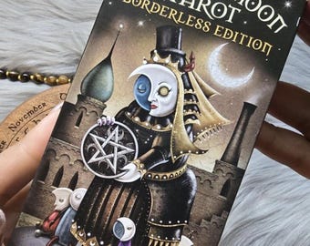 May include: A tarot card deck box with the title "Deviant Moon Tarot Borderless Edition" in gold lettering. The box features a gothic illustration of a figure with a moon face, holding a pentagram, and surrounded by other figures.