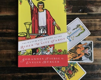Könnte beinhalten: Ein Buch mit dem Titel "THE ULTIMATE GUIDE TO THE RIDER WAITE TAROT" mit gelbem Einband und einer Illustration einer Figur in roter Robe. Mehrere Tarotkarten sind neben dem Buch ausgebreitet, mit Bildern von Schwertern, einer gehörnten Figur und einem Pentakel.