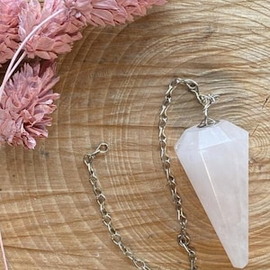 Op de afbeelding: Een roze kwarts pendel, lichtroze en facetgeslepen, met een zilveren ketting en sluiting. De kristal hangt aan een zilveren ketting. Gedroogde roze bloemen staan op de achtergrond, op een houten oppervlak.