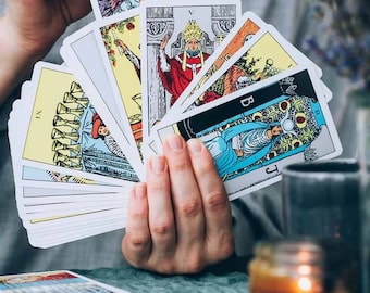 Könnte beinhalten: Eine Hand, die ein aufgefächertes Tarotkartenset hält, jede Karte mit farbenfrohen Illustrationen von Figuren und Symbolen. Die Karten werden über einer türkisfarbenen Oberfläche gehalten, mit einer brennenden Kerze und anderen Objekten im Hintergrund. Wahrsagekarten.