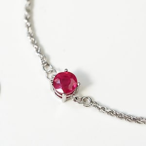 May include: A silver-toned chain bracelet featuring a single round, faceted, deep pink gemstone set in a four-prong metal setting. The bracelet includes a delicate chain with an adjustable extension ending in a small heart-shaped charm stamped 925.