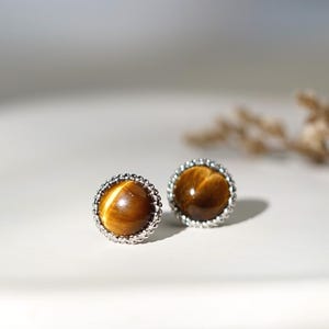 May include: A pair of round stud earrings with a polished, golden-brown tiger's eye stone set in a silver-colored, beaded frame. The earrings are displayed on a white surface with soft lighting.